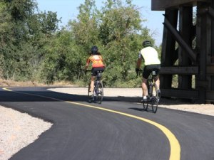 Two cyclists riding bicycles on a paved path curving beneath a wooden bridge, surrounded by trees.