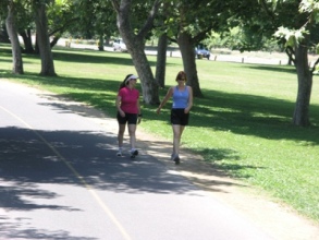 Two women walking on a paved path in a park, under the shade of trees, on a sunny day. The woman on the left wears a pink shirt, black shorts, and a visor, while the woman on the right wears a blue tank top and black shorts.