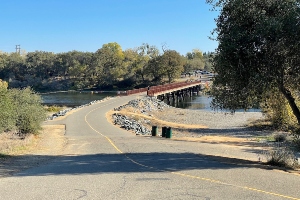 A paved pathway slopes down towards a river, leading to a long brown pedestrian bridge extending across the water; gravelly banks and trees line the river and bridge sides under a clear blue sky.