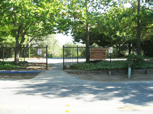 Open wrought-iron gate leading into a park, surrounded by lush green trees, with signs posted on either side of the entrance and a paved pathway.
