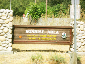 Wooden sign with text "SUNRISE AREA AMERICAN RIVER PARKWAY COUNTY OF SACRAMENTO", set between stone pillars, with a small park logo, vegetation in the background.
