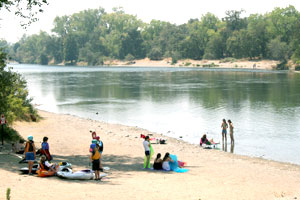 People relaxing on a sandy riverbank with kayaks and inflatables, facing a calm, wide river with trees lining the far bank under a hazy sky.