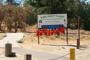 Sign with the text "KIDS DON'T FLOAT - Give Them Something That Will" and a Life Vest Loan Program, displaying several orange life vests hanging on pegs next to a paved path and a water fountain at a park.