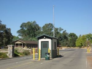 Security gatehouse with a closed green door and small window, a tall silver antenna, and a green trash can, located at the entrance to a road with a residential building visible in the background against a blue sky.