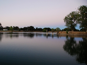 Still water lake reflects a dusky sky and trees along the shore, with a small bridge spanning across the center.