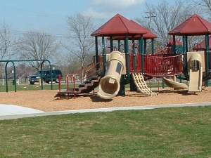 Playground with tan slides, climbing structures, and red accents, set on a wood chip base with a grassy area in front.