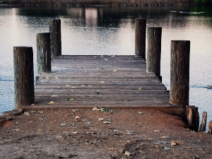 Wooden pier extending into calm water, supported by thick wooden pilings, with a worn, weathered surface and reflections on the water.