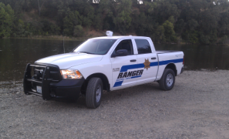 White and blue "RANGER" marked vehicle parked on a gravel beach next to a river with trees in the background.
