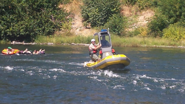 A park ranger in a small yellow and grey rescue boat with a blue light on top navigates a river, looking towards three people floating in inner tubes in the background.